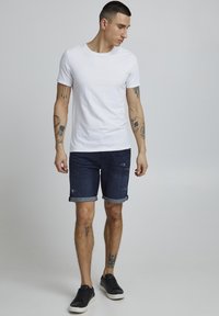 Camiseta blanca de manga corta combinada con pantalones cortos de mezclilla azul oscuro con dobladillos enrollados. Zapatillas negras completan el look casual.