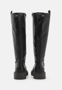 Botas de cuero negro hasta la rodilla con un acabado suave, suelas texturizadas y una marca dorada en la parte trasera. Cuentan con una cremallera para facilitar su uso.