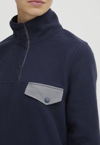 Pullover de forro polar azul marino con un cuello alto, bolsillo de solapa gris con botones y un acento gris claro a lo largo de la tapeta. Textura suave, diseño deportivo.