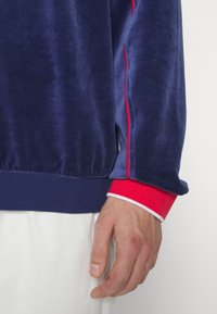 Námořnická modrá sametová mikina s červenými bočními pruhy, žebrované manžety v červené a bílé barvě. Měkká textura, uvolněný střih a elastický pas.