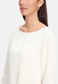 Weiße, leichte Bluse mit rundem Halsausschnitt und langen, leicht ausgestelltend Ärmeln. Glatte Textur und subtile Nähdetails sind sichtbar.