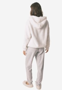 Biała polarowa bluza z kapturem, zestawiona z jasnoszarymi spodniami dresowymi o subtelnym wzorze. Model ma na sobie beżowe kapcie.