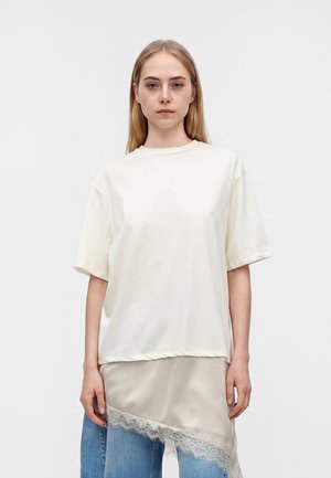 Jeune femme aux cheveux blonds et raides portant un haut blanc à manches courtes oversize superposé à une jupe en satin avec bordure en dentelle et un jean bleu.