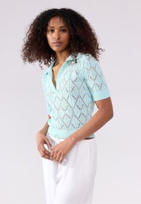 Polo corto a maniche corte di colore verde menta lavorato a maglia, con colletto aperto e motivo a rombi, abbinato a pantaloni bianchi. Materiale leggero e strutturato.