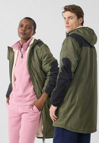 Dos modelos llevan chaquetas: una en verde oliva con detalles en negro, y la otra en rosa claro. Ambas chaquetas cuentan con capucha y cremallera.