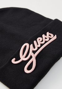 Juodas megztinis kepurė su rožiniu siuvinėtu "Guess" logotipu ant krašto, išsiskirianti lygia tekstūra ir minimalistiniu dizainu.