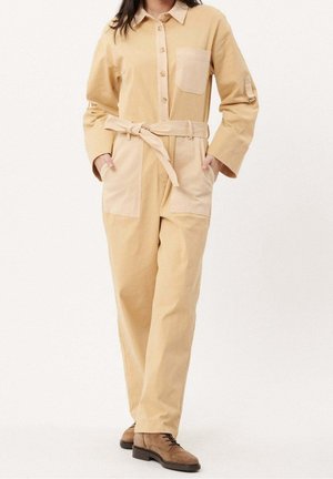 Femme portant une combinaison beige boutonnée avec ceinture à nouer, grandes poches avant et poitrine, associée à des chaussures en daim marron à lacets.