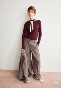 Bordeauxkleurig langemouwtop met witte koordjes en oversized grijze textuurbroek. Model houdt een gedeelte van de broek vast.