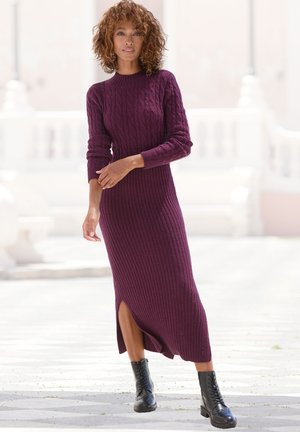 Strickkleid in tiefem Lila mit gerippter Textur, langen Ärmeln und seitlichem Schlitz. Getragen mit schwarzen Schnürkampfstiefeln.