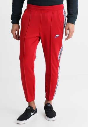 Mężczyzna noszący jaskrawoczerwone spodnie dresowe Nike z białymi i niebieskimi paskami po bokach oraz czarne buty Nike, stojący na białym tle.