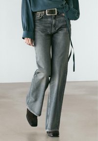 Šedé džínsy s širokými nohavicemi s vybledlým efektem, klasickým pětikapsovým designem a prošitými detaily. Nošeny s tmavým pásem a vrškem.