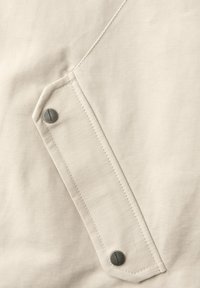 Beige stof med en rektangulær lommeklap, sikret med to metal trykknapper mærket "EASTCORE."