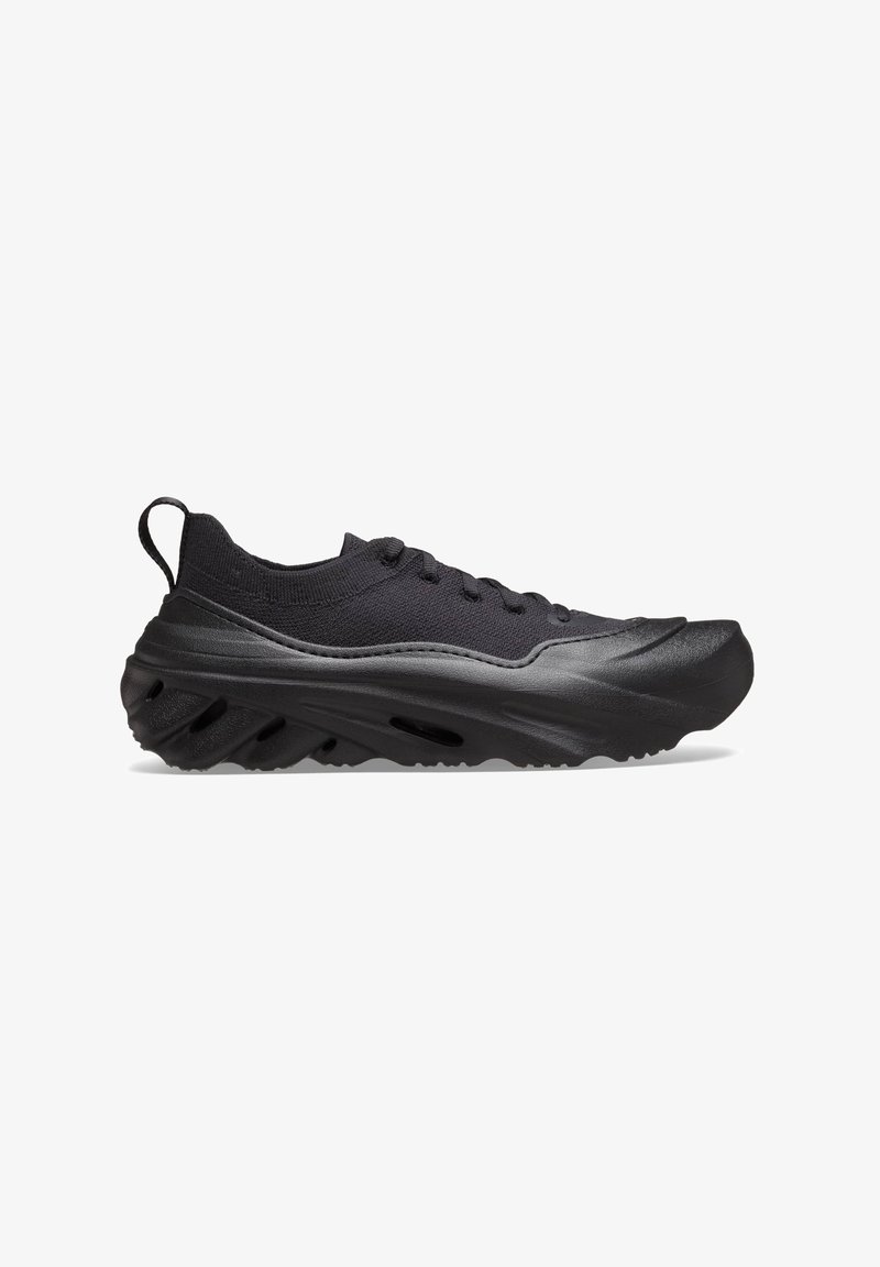 Sneaker sportivo nero con suola spessa e texture, parte superiore in maglia, design con lacci e linguetta sul tallone, mostrato in profilo laterale su sfondo bianco.