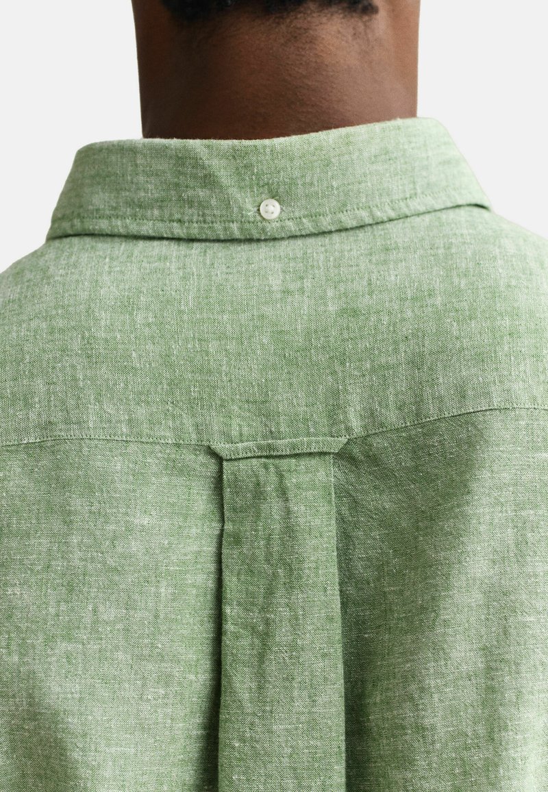 Vista posterior de una persona que lleva una camisa de lino verde claro con una trabilla de cuello abotonada y un pliegue central en la parte trasera.