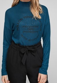 Teal långärmad topp med hög krage och grafisk text som har rhinestones, kombinerad med svarta högmidjade byxor och tygbälte.