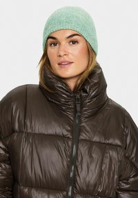 Chaqueta acolchada marrón con cuello alto y cierre de cremallera, combinada con un gorro de punto acanalado en verde claro. Textura suave y material ligero.