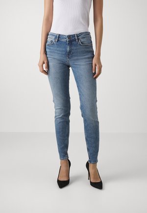 Mujer vestida con vaqueros ajustados azul claro, top sin mangas blanco y zapatos de tacón altos negros, de pie frente a un fondo liso.