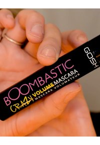 Svart mascaraflaska som hålls i en hand, märkt "BOOMBASTIC CRAZY VOLUME MASCARA" i rosa och gula bokstäver. Slät yta, rektangulär form.
