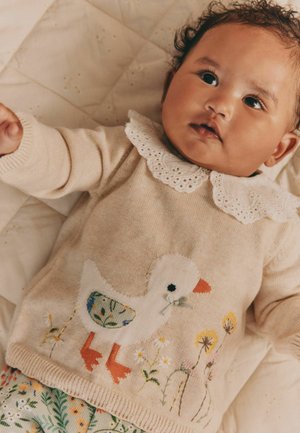 Säugling mit lockigem Haar liegt auf einer gesteppte Decke, trägt einen beigen Pullover mit weißen Enten und Blumen sowie eine floral gemusterte Hose.