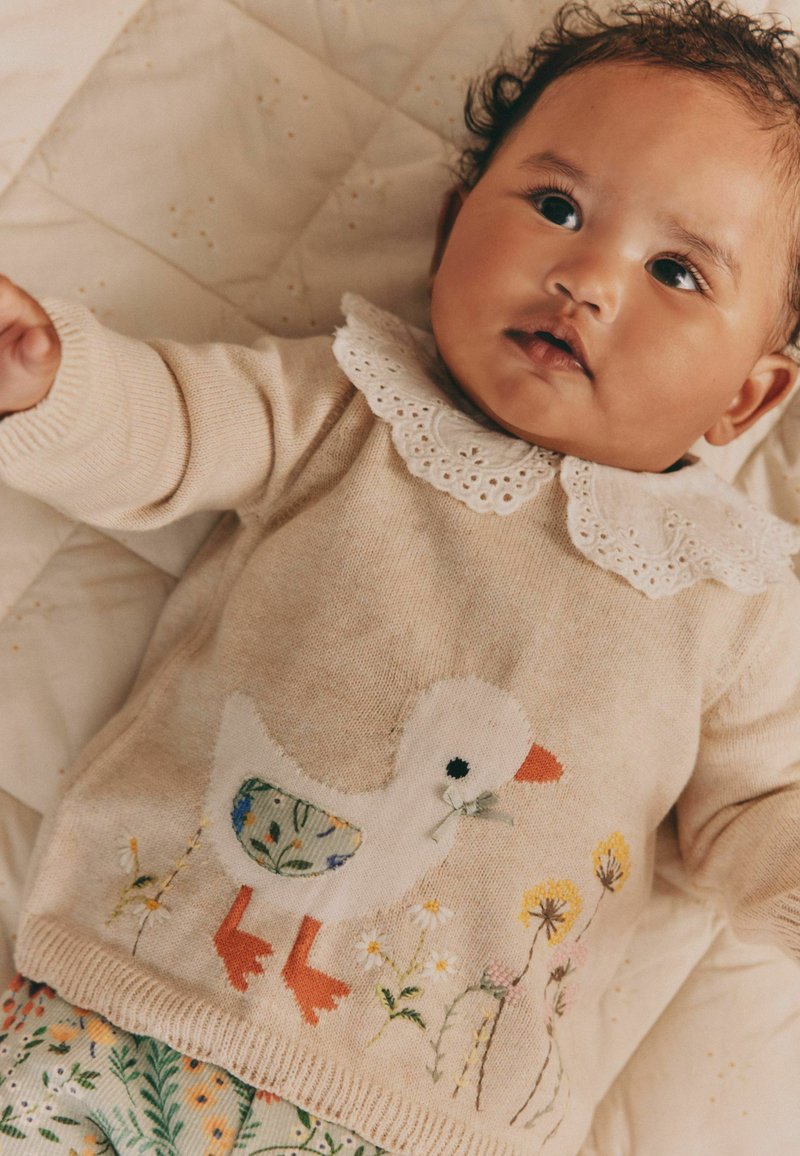 Spædbarn med krøllet hår ligger på en quiltet tæppe iført en beige sweater med en hvid and og blomster, samt bukser med blomstermønster.