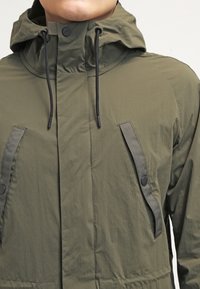 Uomo che indossa una giacca a cappuccio verde oliva con stringhe nere e tasche a pattina verticali fissate da bottoni a pressione neri.