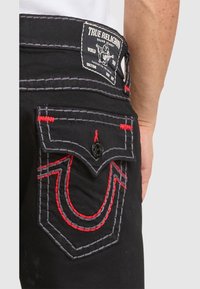 Pantaloni True Religion negri, cu detalii de cusătură roșie și gri pe buzunar și un patch de marcă vizibil pe talie, purtați de o persoană.