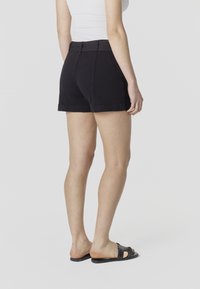 Femme portant un short noir taille haute et des sandales à glissière noires cloutées, debout devant un fond clair uni, vue de dos.