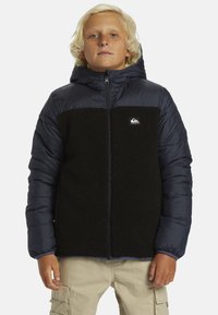 Blond chlapec v čiernom flísovom saku a námorníckom pufrovom kabáte s kapucňou, a béžových cargo nohaviciach, stojí s neutrálnym výrazom.
