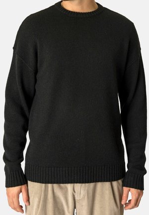 Mand iført en ensfarvet sort strikket sweater med ribkant ved ærmer og bund, kombineret med lyse beige fløjlsbukser.