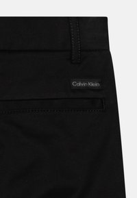 Juodi kelnių su Calvin Klein etikete, lygia tekstūra, standartine juosta ir subtiliu siuvimo detaliu šalia kišenės.