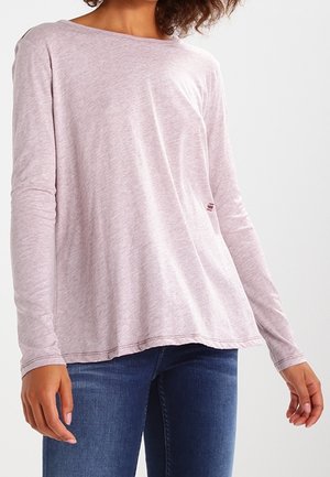 Kvinde iført en lyserød langærmet skjorte og blå jeans, stående mod en ensartet lys baggrund.