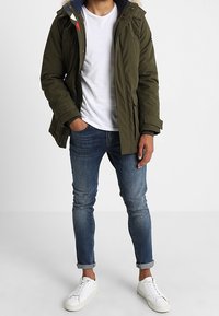 Olivgrön parkas med huva, framfickor och dragkedja. Kombination med åtsittande blå jeans och vita sneakers. Enkelt, funktionellt design.