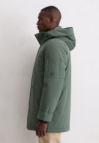 Zijaanzicht van een persoon in een groene winterjas met capuchon en beige broek tegen een eenvoudige witte achtergrond.
