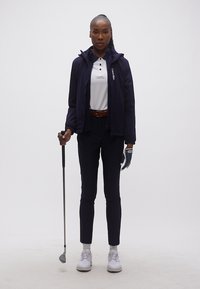 Γυναίκα που στέκεται κρατώντας ένα μπαστούνι γκολφ στο δεξί χέρι, φορώντας μπλε σκούρο σακάκι, λευκό πόλο, μαύρο παντελόνι, γάντι γκολφ και λευκά αθλητικά παπούτσια.