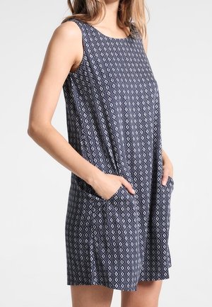 Vestido azul marino sin mangas con patrones de diamantes blancos, llevado por una persona con las manos en los bolsillos laterales, de pie frente a un fondo liso.