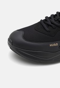 Chaussure de sport noire avec des matériaux en mesh et lisses, design à lacets, semelle épaisse et marquage "HUGO" sur le côté.