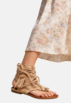 Pied portant une sandale à lacets en daim beige perforé avec des pompons, associée à une jupe midi légère à fleurs.