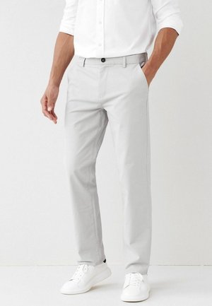 Mężczyzna w jasnoszarych spodniach, białej koszuli na guziki z podwiniętymi rękawami oraz białych trampkach, stojący z jedną ręką w kieszeni.
