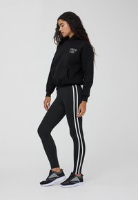 Felpa nera con tasca frontale e testo bianco, abbinata a leggins neri con strisce bianche sui lati. Modello indossa scarpe sportive nere.