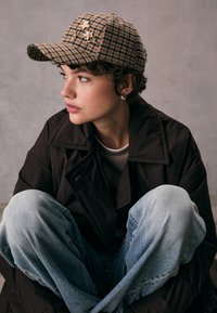 Cappellino marrone a quadretti con ricamo, abbinato a un cappotto oversize scuro e jeans azzurri chiari. Il cappellino presenta una visiera curva e un tessuto strutturato.