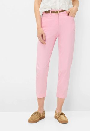 Frau trägt hellrosa kurze Hose, braunen Gürtel, weiße Spitzenbluse und beige Freizeitschuhe, steht mit einer Hand in der Tasche.