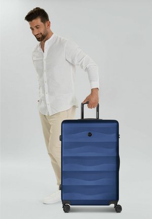 Mann in weißem Hemd und beiger Hose steht und hält den Griff eines großen blauen Rollkoffers mit strukturierter Oberfläche.