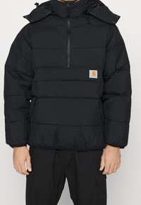 Svart puffjacka med hög krage, halvdragkedja framtill, påfällsficka och elastiska muddar. Har en logotypmärke på bröstet. Isolerad textur.