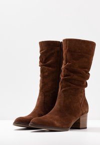 Botas de tobillo de ante marrón con un tacón apilado, que presentan un diseño fruncido y un zipper lateral. Textura suave y punta redondeada.