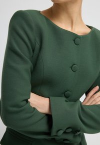Grüne strukturierte Jacke mit runder Halslinie, langen Ärmeln mit breiten Bündchen und auffälligen runden Knöpfen an der Vorderseite. Glatte Textur.
