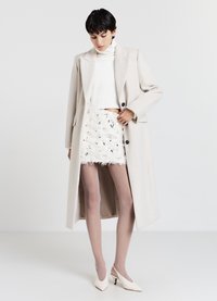 Lungo cappotto beige sopra un dolcevita bianco a coste e una gonna bianca testurizzata con frange. Tacchi beige. Superfici lisce, silhouette sagomata.