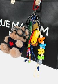 Czarna torba z białym napisem, z pluszowym breloczkiem w kształcie misia noszącym okulary, kolorowymi plastikowymi charmsami oraz różnorodnymi brelokami do kluczy przypiętymi.