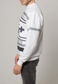 Bílá mikina s černými horizontálními pruhy a geometrickými vzory. Má kulatý výstřih a žebrované manžety, doplněná šedými džínami.