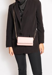 Borsa rettangolare rosa realizzata in pelle liscia con chiusura a pattina, tracolla in catena dorata e design minimalista. Indossata a tracolla.