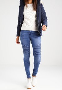 Frau trägt eine marineblaue Jacke, einen weißen strukturierten Pullover, enge blaue Ripp-Jeans und weiße Sneaker und steht vor einem einfarbigen hellen Hintergrund.